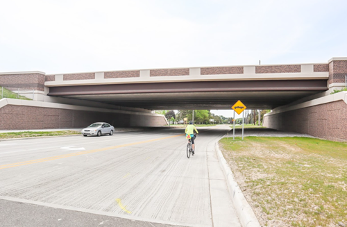 Car and cyclist on roadway with bridge overhead in daylight.