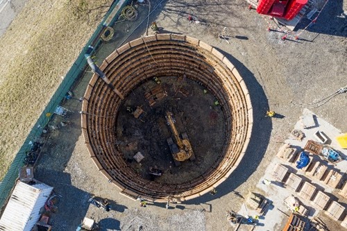 Aerial view into the tunnel shaft. Excavator with wooden planks line the tunnel shaft for saftey and support. Workers can also be seen at the bottom of the tunnel.